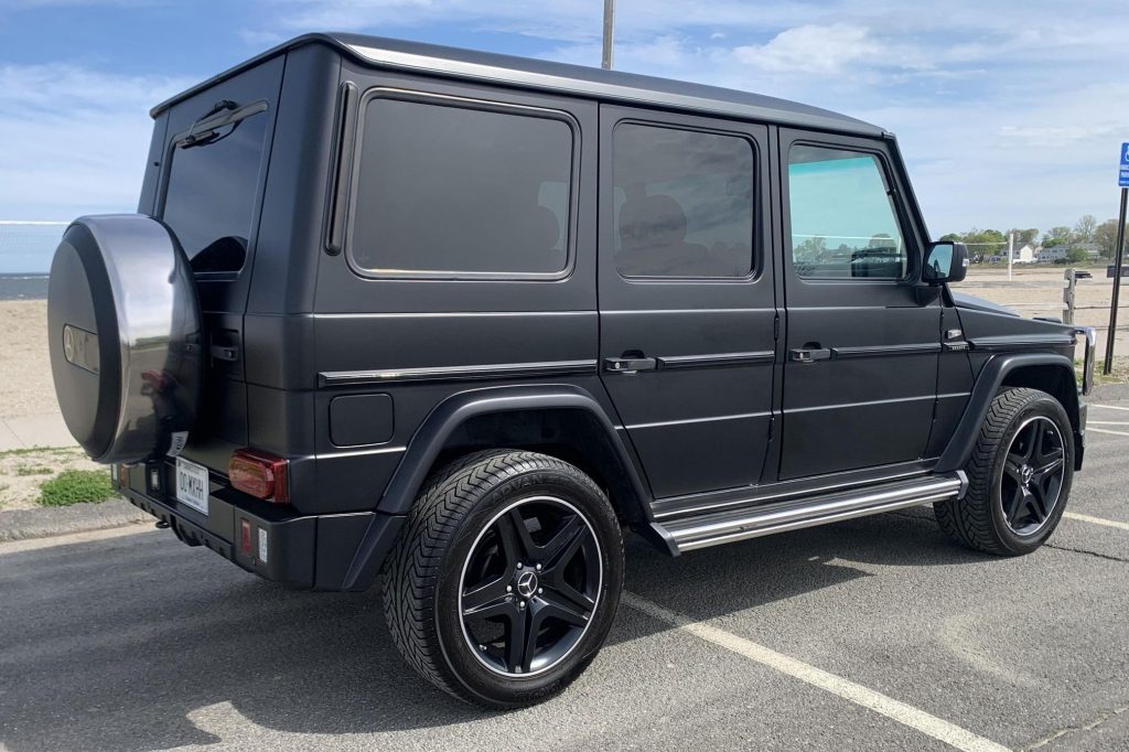 The rear 3/4 view of a modified matte-black 1995 Mercedes-Benz G320 on a beachside parking lot