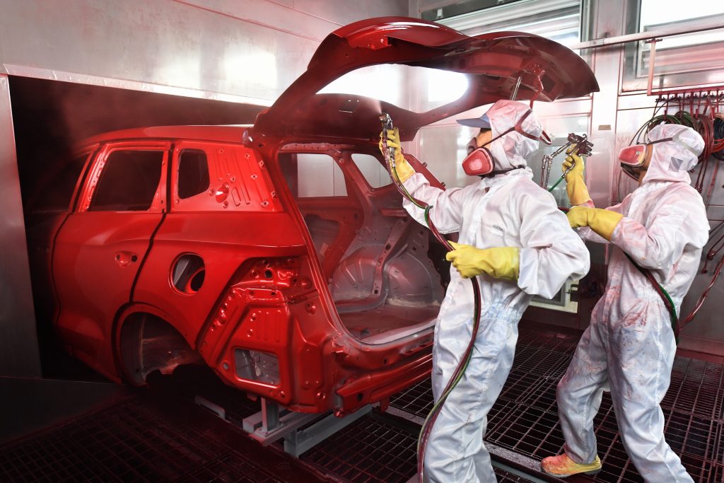Technicians in full-body protective gear and respirators spray paint the body of a car red