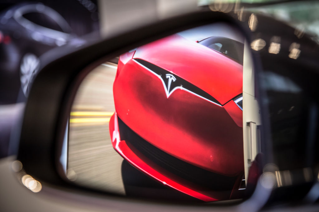 A red Tesla is shown approaching through another car's wing mirror