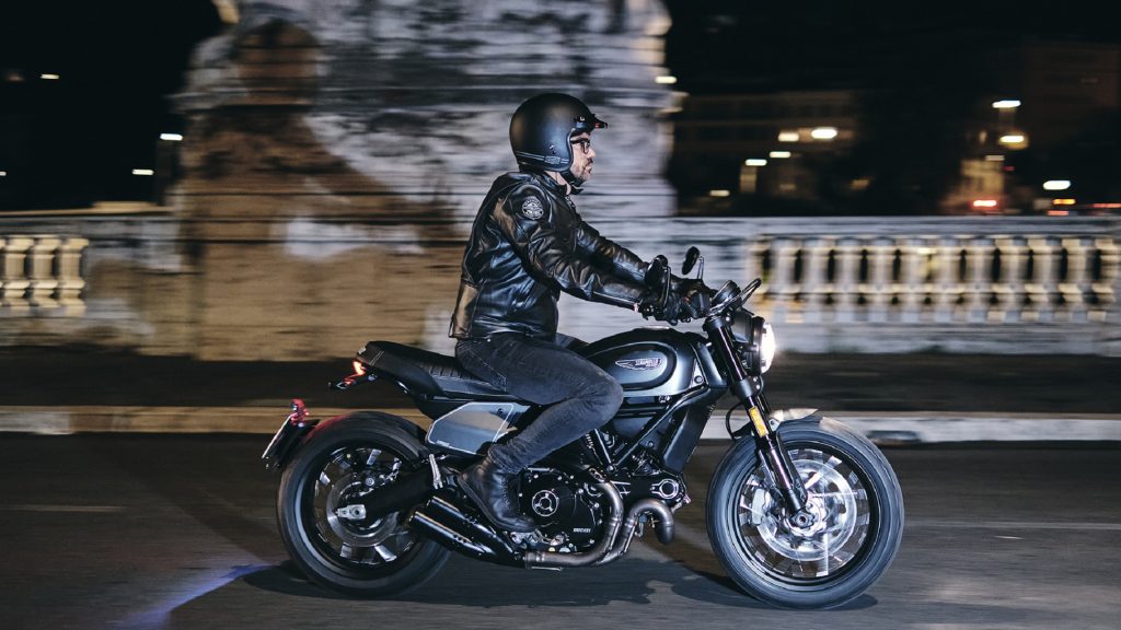 A black-clad rider on a gray-and-black 2021 Ducati Scrambler Nightshift riding through a city at night